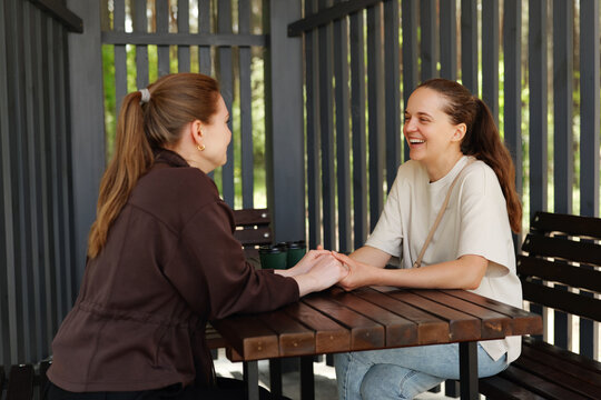 Two friends sit at a wooden table in a garden. They hold hands and chat happily. Sunlight comes through the trees around them
