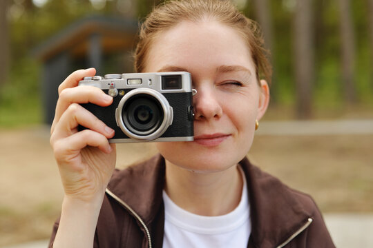 A woman stands in a park holding a vintage camera up to her face. She looks through the viewfinder while squinting her eyes. Trees surround the area