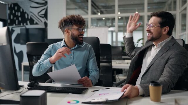 Two successful multiethnic business men team comparing data in documents and computer celebrate giving high five partnership success result diversity businessmen with paperwork job growth in office