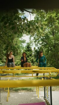 Three women are seen relaxing and talking on a yellow monkey bar structure outdoors. The friends enjoy a casual moment on playground equipment in a green park environment