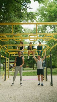 A group of friends enjoys an outdoor calisthenics workout at a park. Two men exercise on monkey bars while three women watch and smile