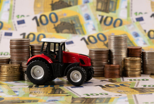 A symbolic miniature tractor and stacks of coins against a background of 100 euro banknotes