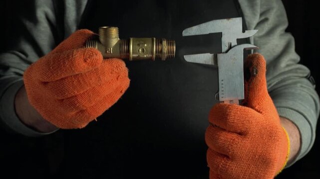 Technician in orange gloves measures brass fitting with caliper tool, showcasing precise measurement in a well-lit workspace with dark background
