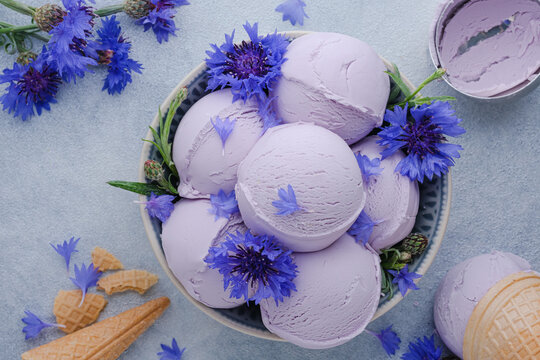 A bowl holds several scoops of lavender ice cream surrounded by blue flowers. Waffle cones sit near the bowl on a light surface. Healthy frozen dessert. top view