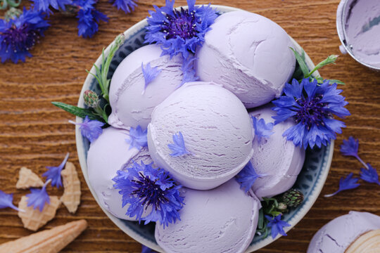 A bowl holds several scoops of lavender ice cream surrounded by blue flowers. Waffle cones sit near the bowl on a wooden background. top view