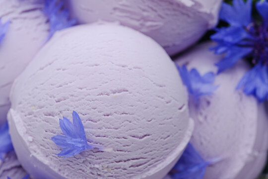 Scoops of lavender ice cream rest next to small blue flowers. The scene captures a dessert display showing the creamy texture and color of the ice cream. This creates an appealing visual treat