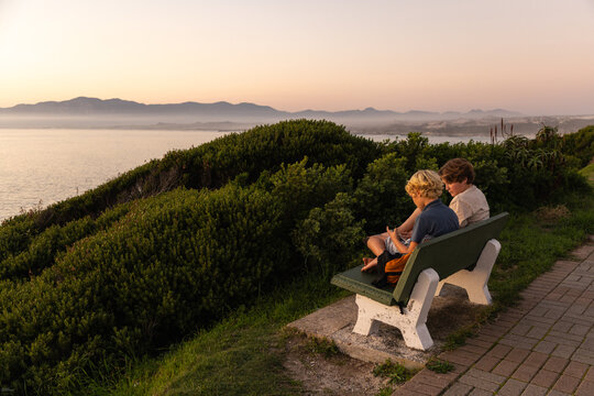 2 pre teen boys sitting on bench overlooking coastline of DeKelders, South Africa