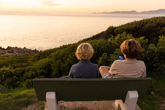 2 pre teen boys sitting on bench overlooking coastline of DeKelders, South Africa