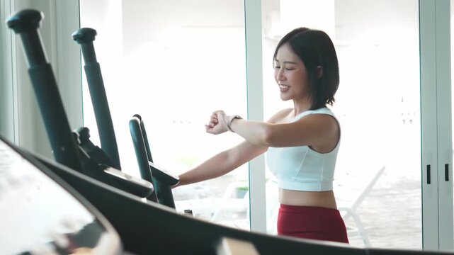 Side view of a happy young Asian woman in sportswear holding the handlebars of a stationary exercise bike during a cardio workout session in a bright gym