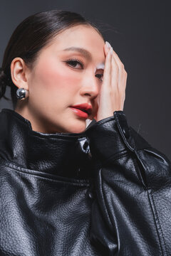 Mysterious Asian woman covering half face in leather jacket for high fashion cosmetics skincare marketing campaign intense eye contact dualism concept edgy portrait studio
