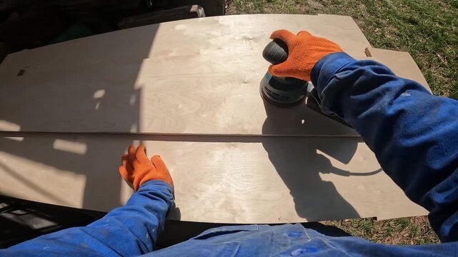 The master sands a wooden surface with an eccentric machine. The process of woodworking. Hands in orange work gloves use an orbital sander to level the plywood. First person view