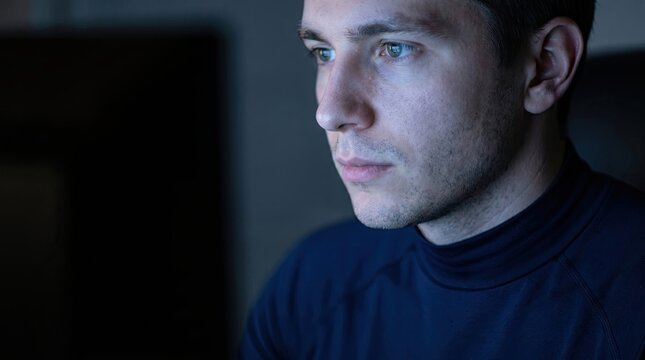 Young man concentrated while staring at glowing computer screen in dark room at night time session.