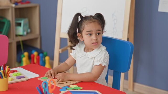 Little girl placing a wooden star into a shape sorter puzzle with her hand at a red table inside a building; quiet concentration.