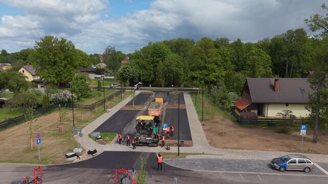Construction crew lays asphalt on new road with heavy machinery and workers in safety vests in a suburban area surrounded by trees