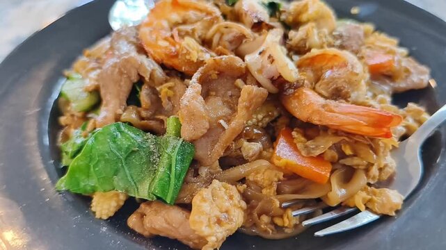 A delicious plate of Pad See Ew with noodles, shrimp, and vegetables. The dish is served on a dark plate with a fork, ready to be enjoyed