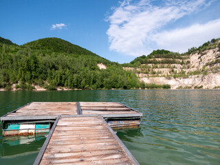 Fototapeta premium Floating wooden dock on a flooded quarry lake in Kralovany, Slovakia. Clear green water, terraced limestone cliffs, forested hills and blue summer sky.