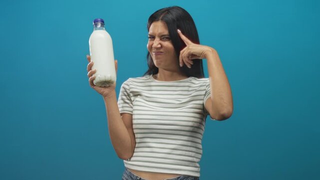 Woman holding milk bottle and pointing finger to temple in studio wearing striped shirt with a wince expression; playful skepticism.