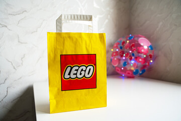 Obraz premium A bright yellow paper bag featuring the iconic red and white LEGO logo stands on a clean white surface. In the soft-focus background, a decorative pink orb with lights adds a playful atmosphere.