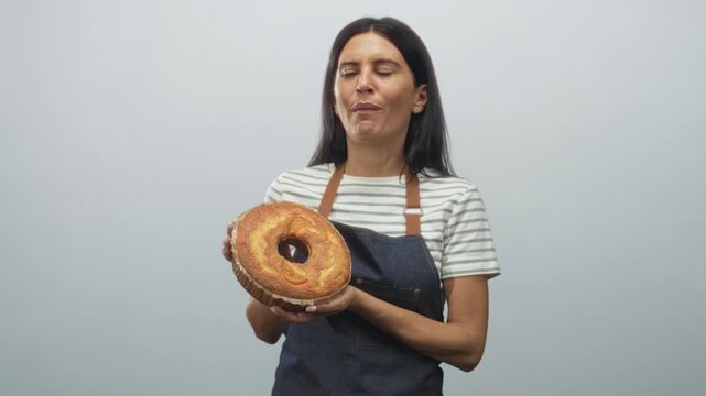 Woman baker holding a ring cake with both hands, wearing an apron over a striped shirt and showing changing expressions from pensive to laughing in studio; joy pride warmth.