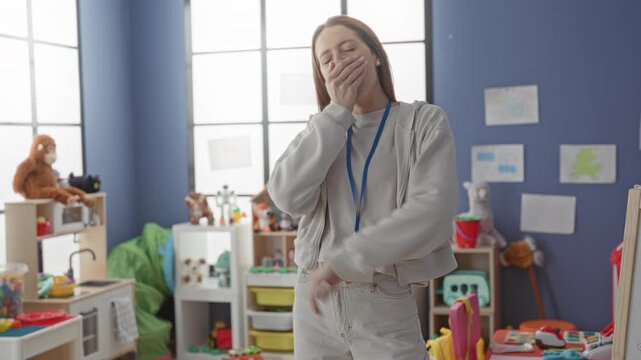 Woman teacher covering mouth while yawning, hand to temple, badge lanyard visible in colorful preschool classroom; fatigue patience.