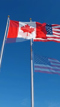 Canadian and American Flags Waving Together in Blue Sky - The Canadian flag and the American flag are shown flying side-by-side on flagpoles against a clear blue sky.