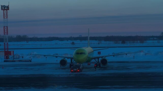 Airplane pushed back by tug on a snow-covered airport tarmac. Ground crew prepares jet for departure in winter twilight at blue hour. Wide shot