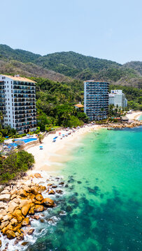 Aerial view of Las Gemelas Beach with turquoise water, white sand, and luxury resorts against lush green mountains and people on the shore in Puerto Vallarta, Jalisco, Mexico.