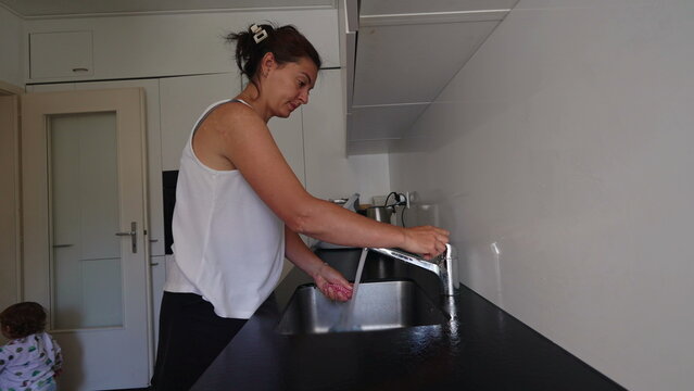 Woman washing kitchen sponge at sink in modern apartment while toddler walks nearby during daily household cleaning routine and family domestic lifestyle