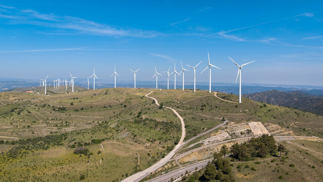Aerial view of large onshore wind farm with multiple turbines stretching across green mountain ridgeline, winding road and agricultural terraces in foreground under clear blue sky