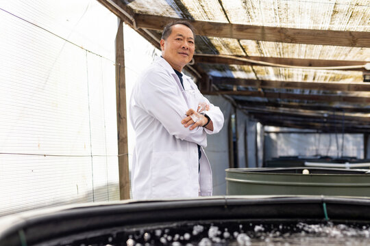 Senior Asian man wearing white lab coat standing arms crossed in hatchery beside aerated tanks