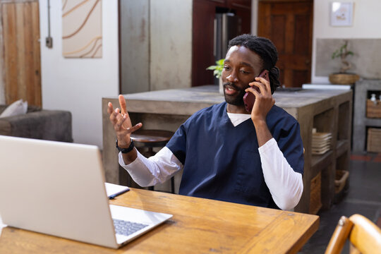 Male medical worker sitting at dining table wearing navy scrubs speaking on smartphone using laptop