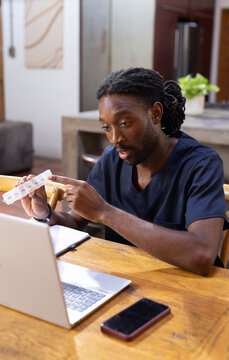African American male wearing navy scrubs holding weekly pill organizer speaking to laptop at home
