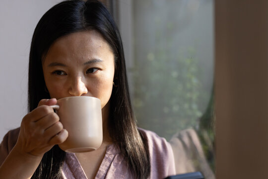 Mature Asian woman in mauve blouse holding beige mug, looking out window in living room, smartphone