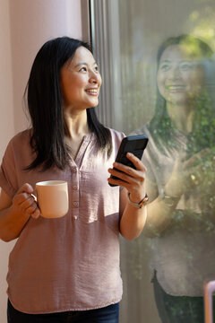 Asian woman standing at home near big window holding mug and phone, wearing dusty rose blouse