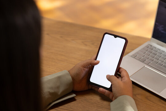 African woman holding phone with blank screen in light blazer, sitting at wood desk with laptop