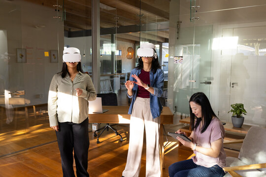 Diverse female coworkers testing VR wearing VR headsets using tablet, laptop in glass office
