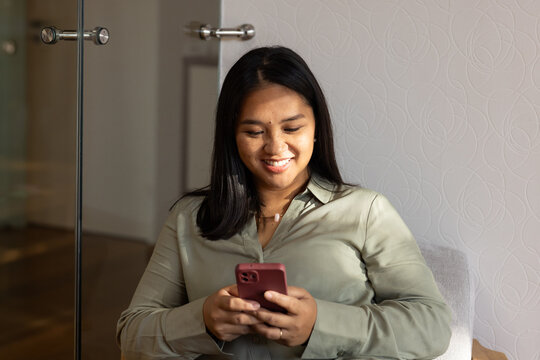 African American woman smiling, holding red smartphone, sitting by glass partition in olive blouse
