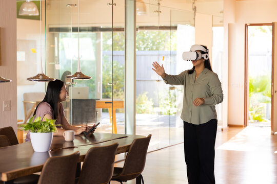 Diverse women coworkers using tablet at table, other wearing white VR headset reaching, copy space