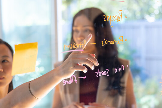Diverse female team collaborating at office pointing at glass whiteboard with yellow note, markers