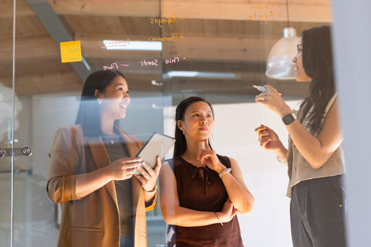 Diverse female team in blazer at office writing on glass board with yellow sticky note, marker