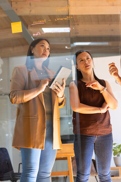 Diverse female pair in business casual standing at office glass board with sticky note, with tablet