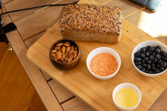 Bamboo cutting board is holding seeded loaf, bowls of almonds, lentils, blueberries on wood table