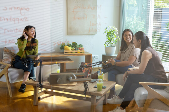 Female friends sitting in living-room office lounge recording podcast with microphones and laptop