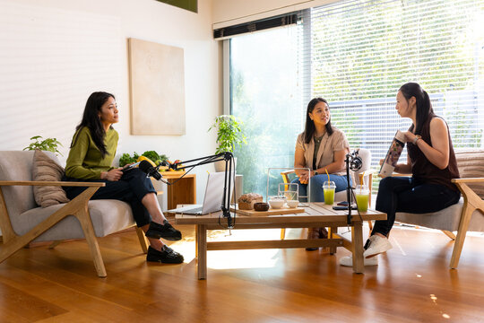 Diverse female colleagues podcasting in living-room around coffee table with mics and smoothies