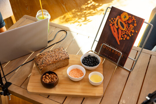 Seeded multigrain loaf is resting on bamboo board on sunlit wooden table, bowls, cookbook, laptop