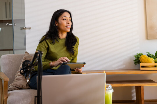 African American adult woman sitting on armchair holding tablet and speaking to microphone at home