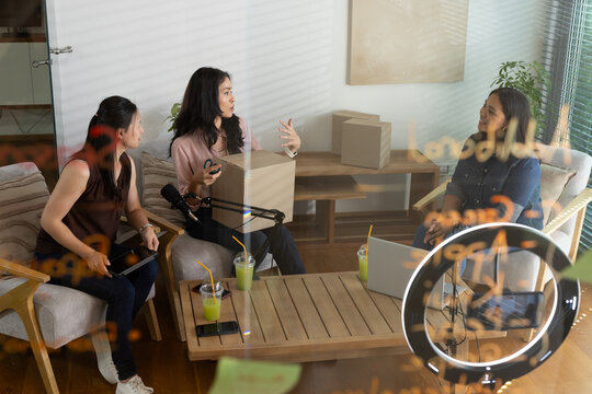 Diverse female creators sitting around coffee table in living room, holding box, using microphone