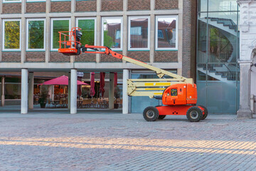An orange articulating boom lift stands on a cobblestone square beside a brick and glass building, with raised work platform, wheels, hydraulic arm, outdoor cafe tables, columns, and reflected windows © Volha