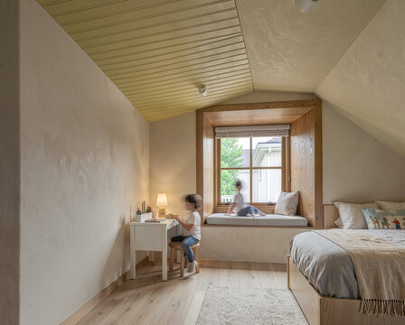 Child studying at wooden desk in cozy attic bedroom with window seat and soft bedding.