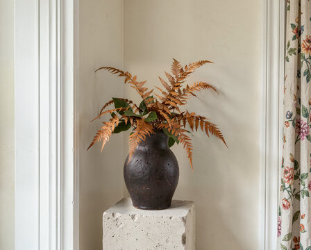 Brown ceramic vase with dried ferns on concrete pedestal in cozy interior corner near floral curtain.